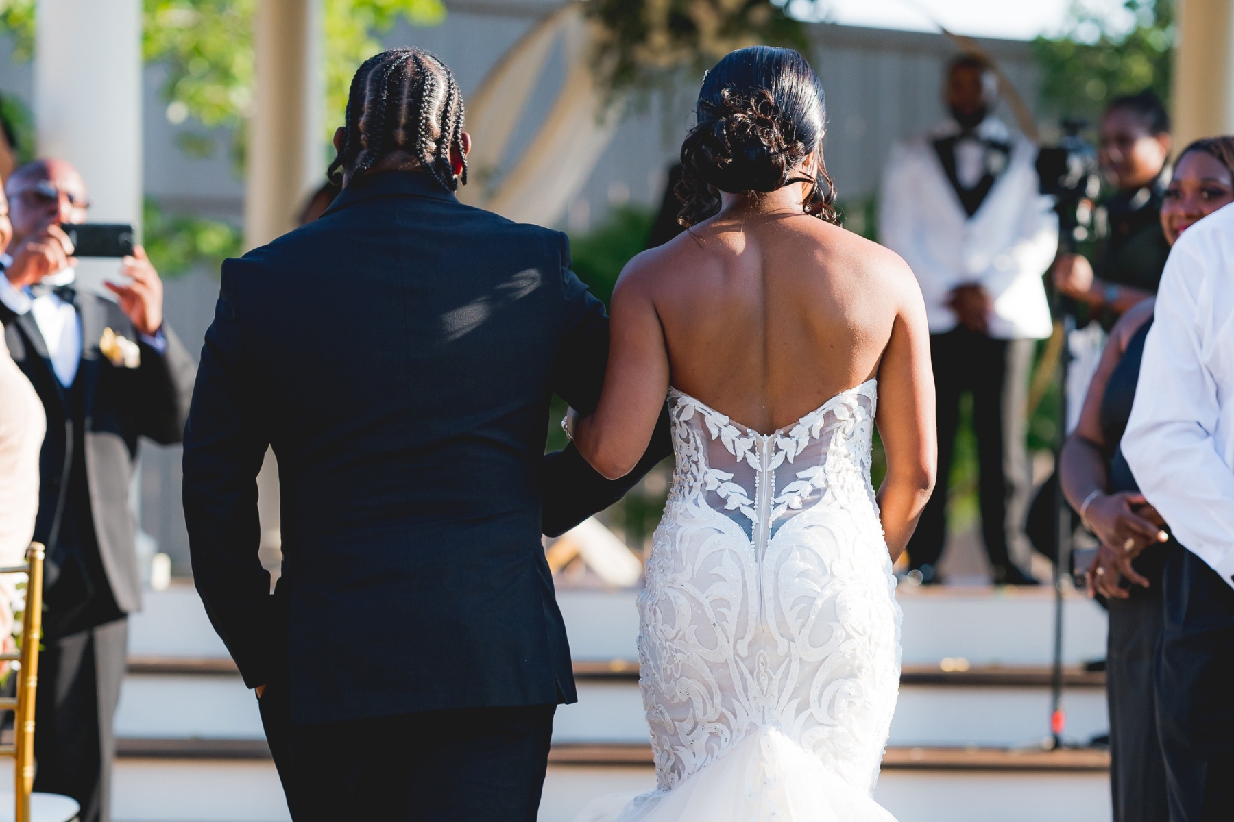 The back of a Black man with cornrows and a Black bride in a strapless wedding gown walking down the aisle