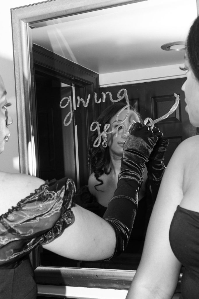 woman writes with lipstick on mirror "giving gems" as another woman looks on