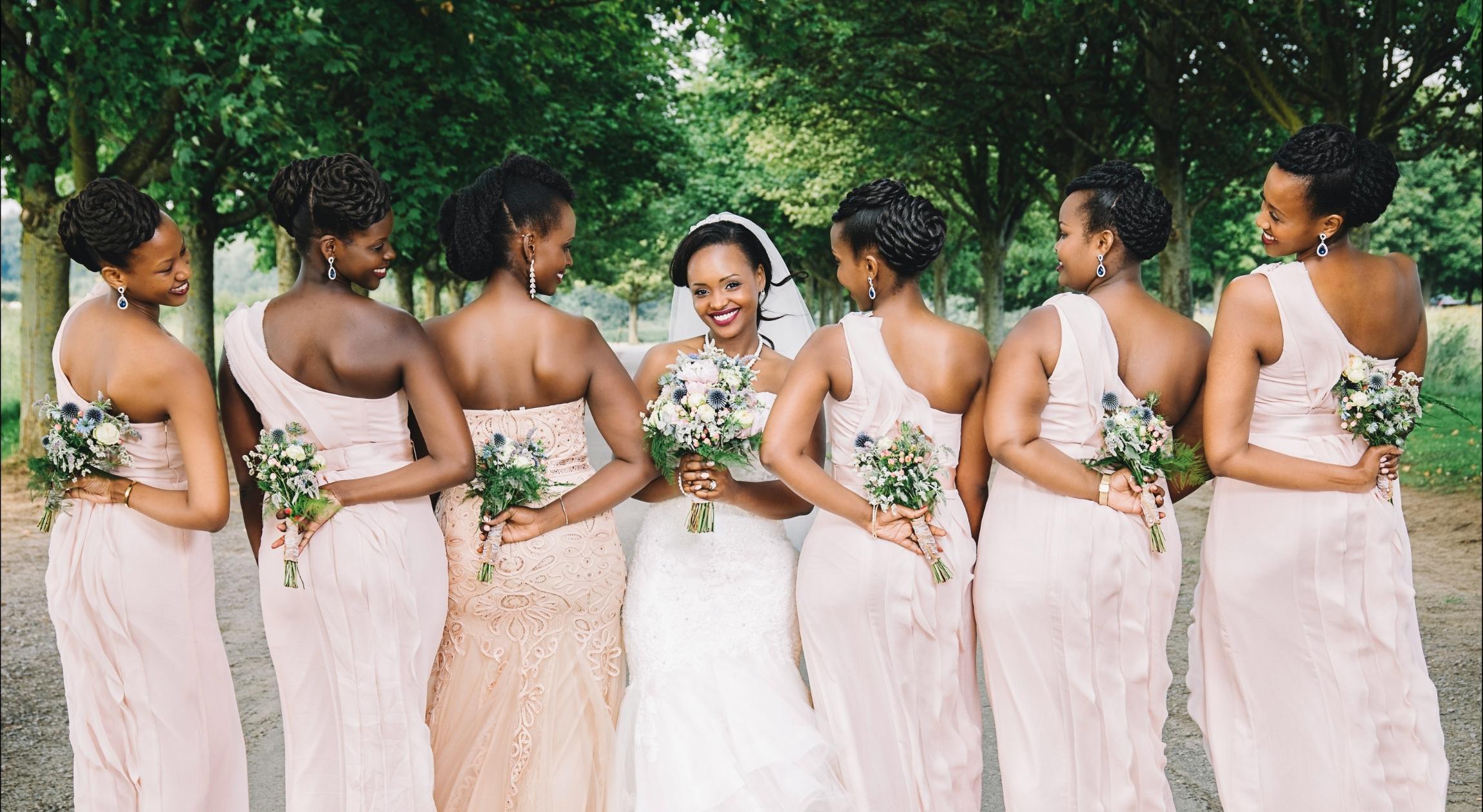 Six Black women in pink dresses stand next to a Black woman in a white dress