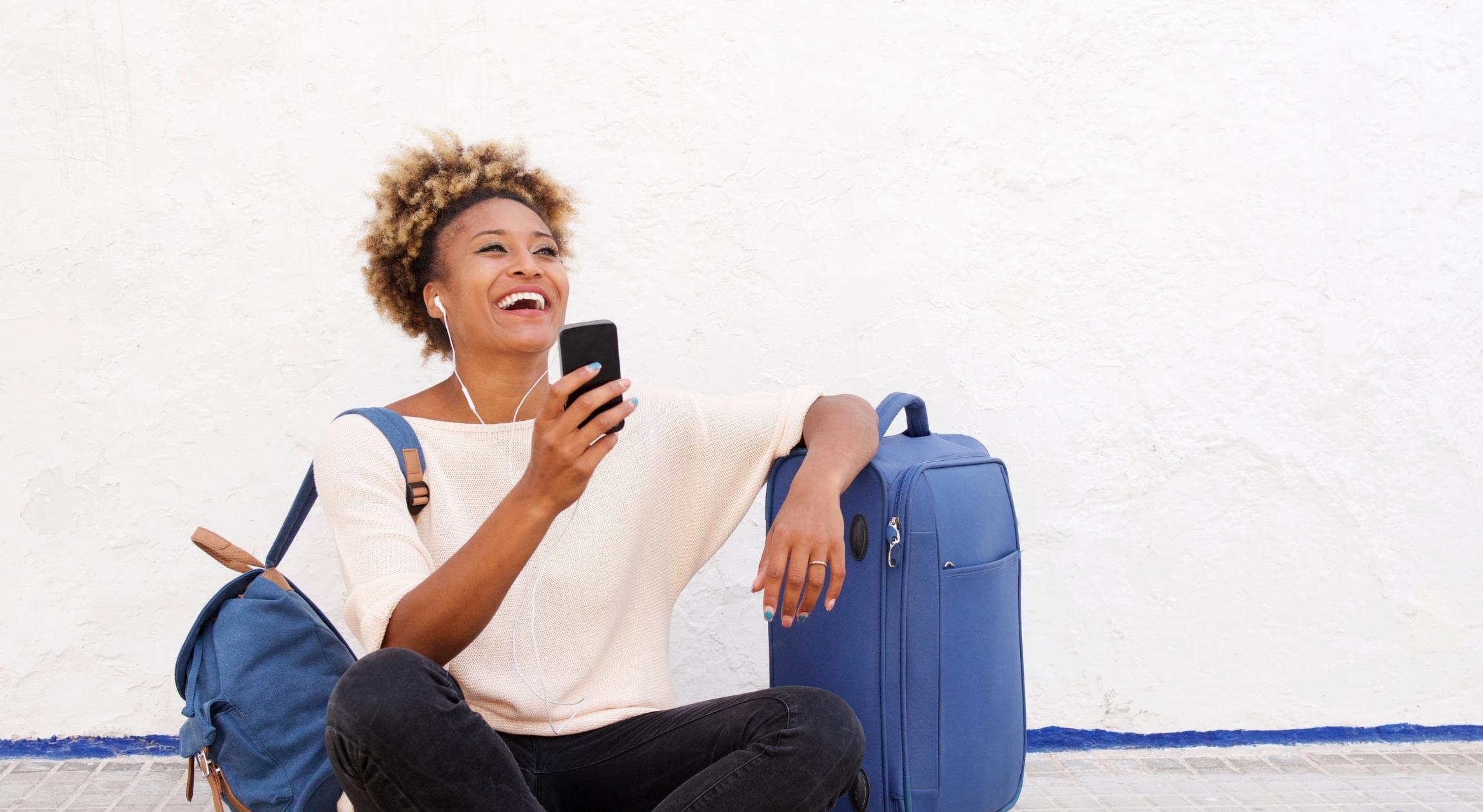 A Black woman sitting on the ground with two pieces of luggage holding on to a cell phone