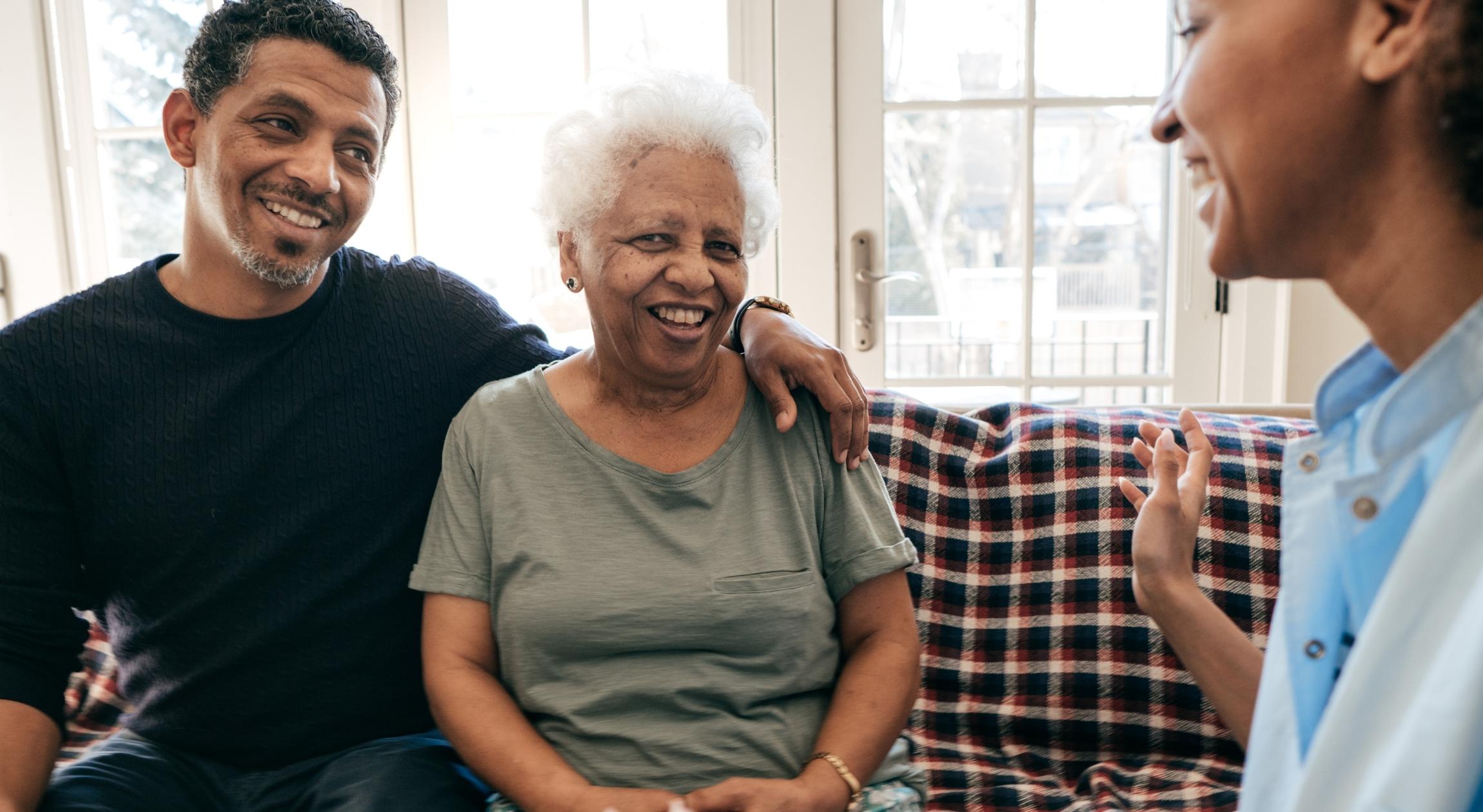 A brown man sits on the couch with his arm over an older brown lady