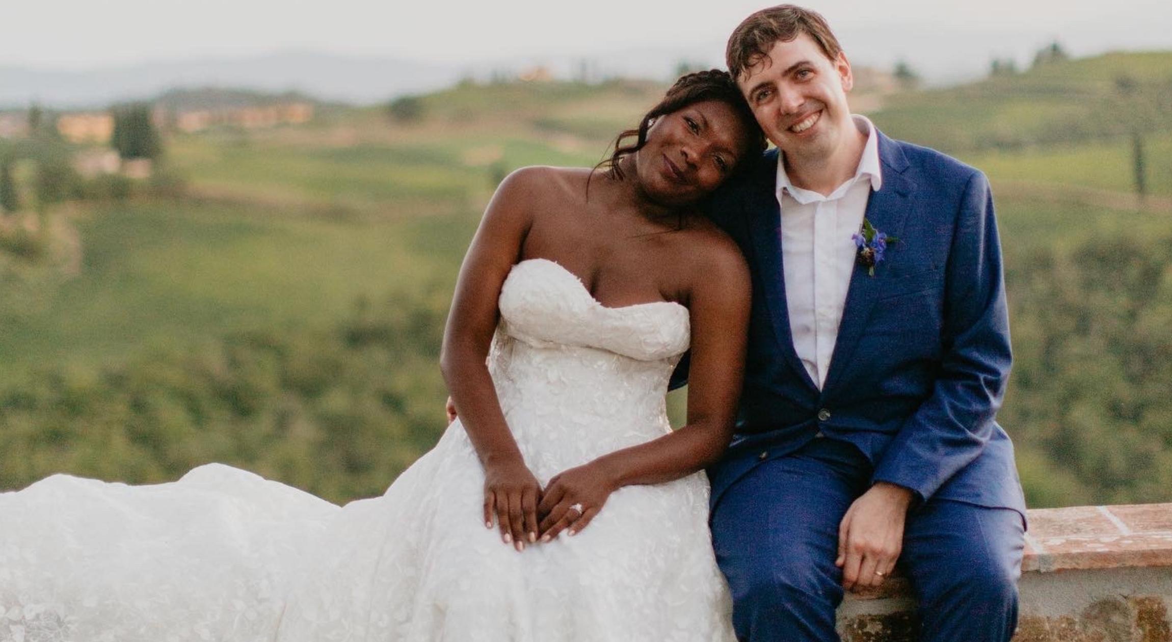 A Black bride sits next to her Italian groom on a ledge in front of the Italian countryside