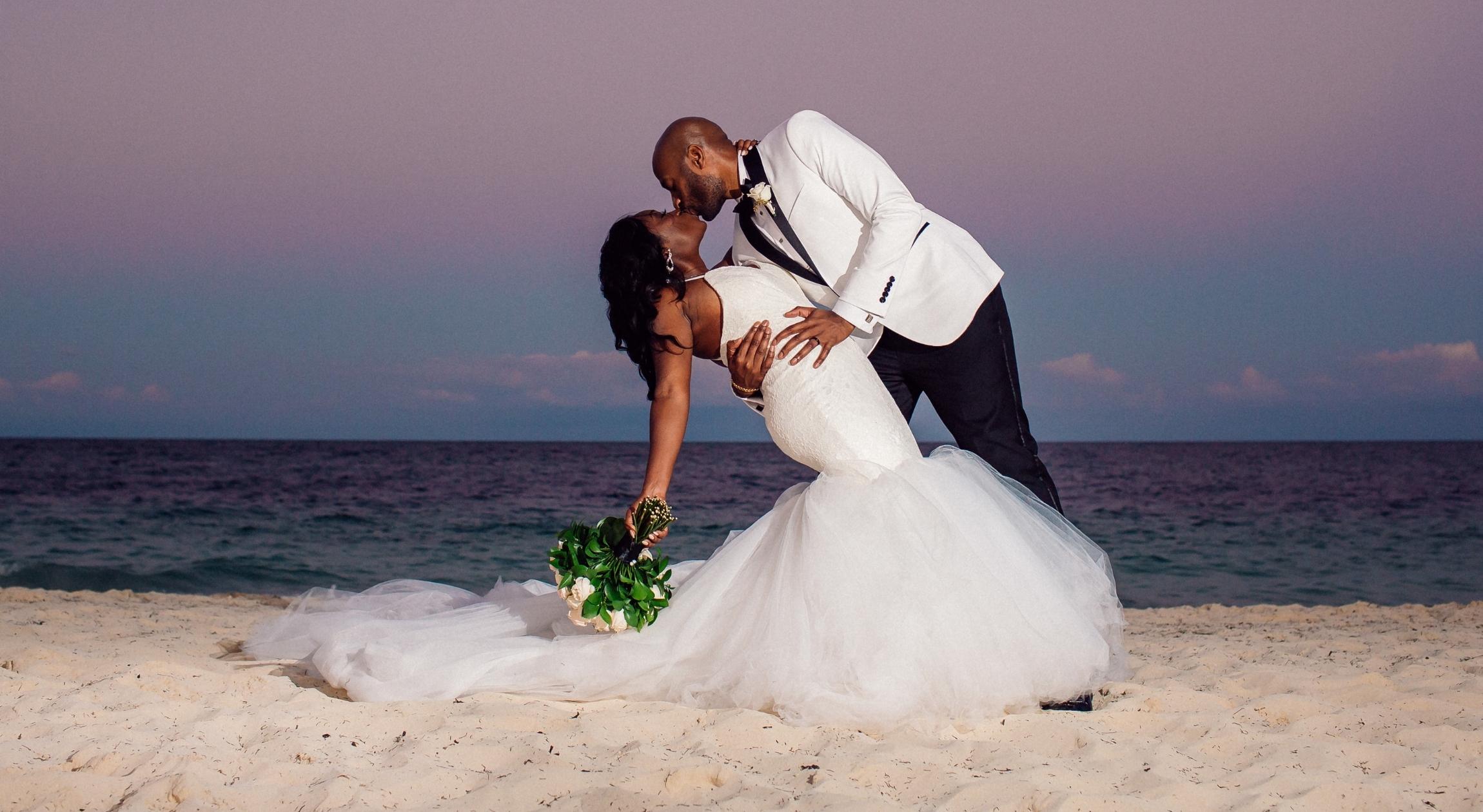A Black couple in wedding attire kissing on the beach