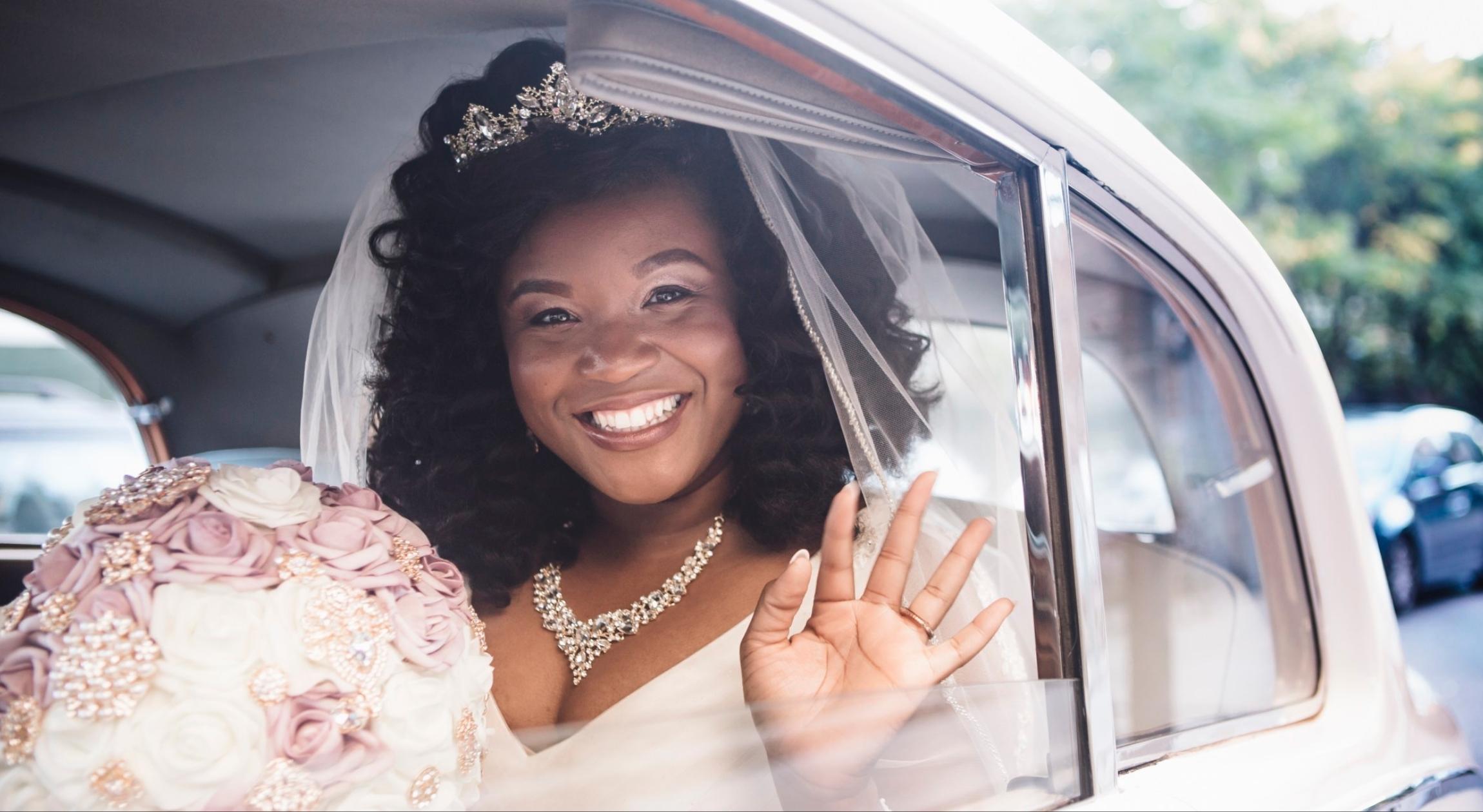 A Black bride in a wedding dress holding a bouquet waving from inside of a car