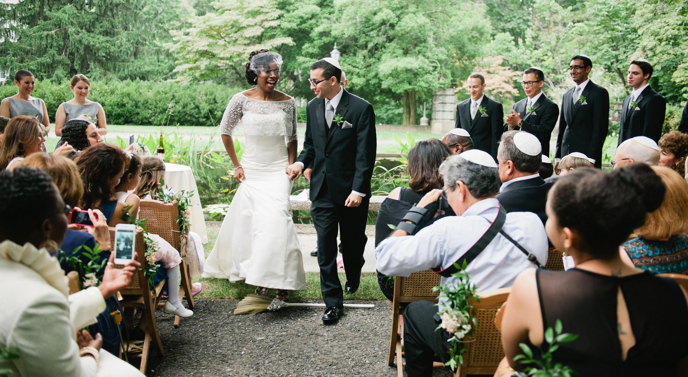 A Black woman and a Jewish man jump over a broom in front of people sitting down