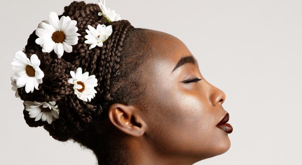 A Black woman with her eyes closed. Her hair is in a braided bun with daisies adorned throughout.