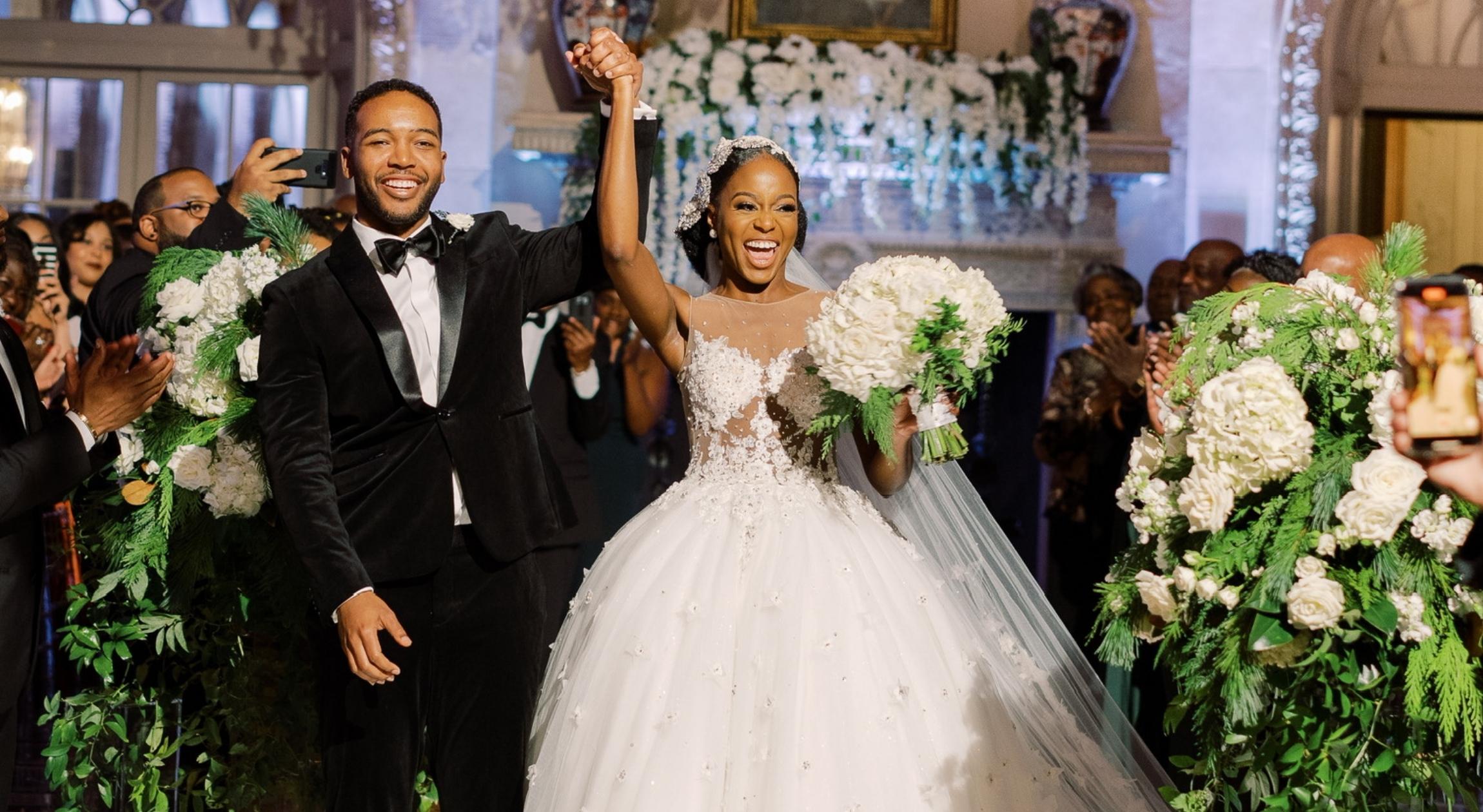 A Black man in a tuxedo holding hands with a Black woman in a wedding dress that is in the ballgown silhouette