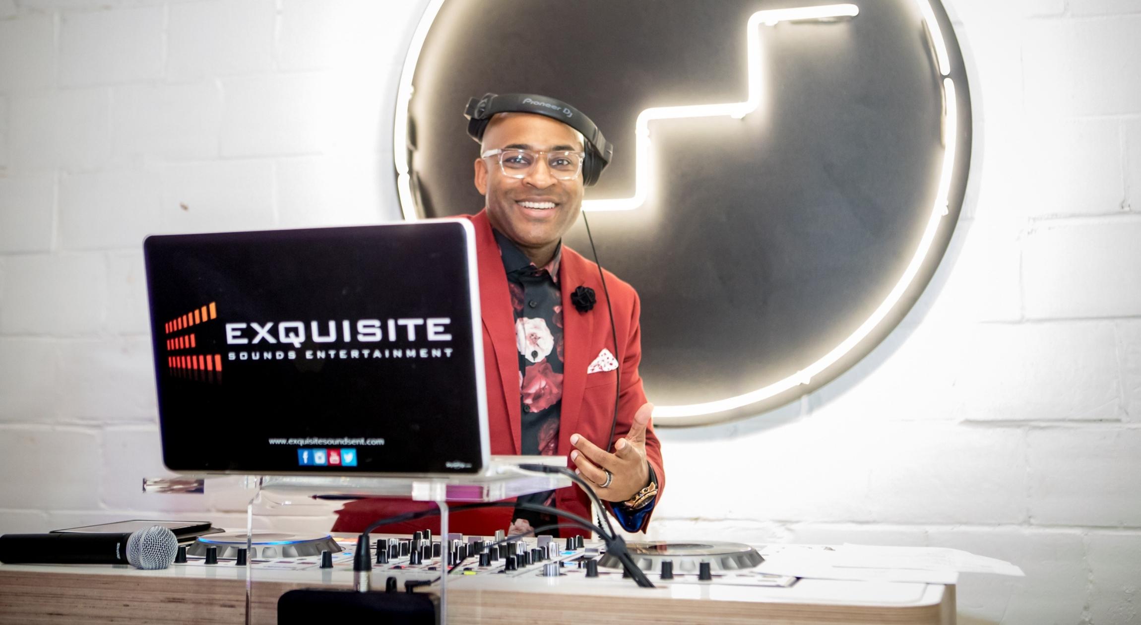 A Black man with headphones on wearing a red suit jacket behind a laptop