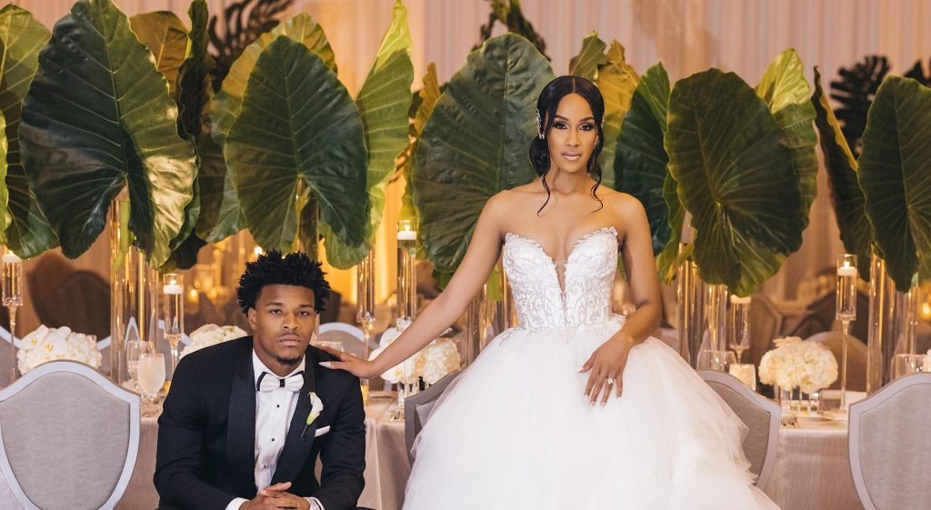 A Black man in a tuxedo sitting down next to a Black woman in a wedding dress that is standing