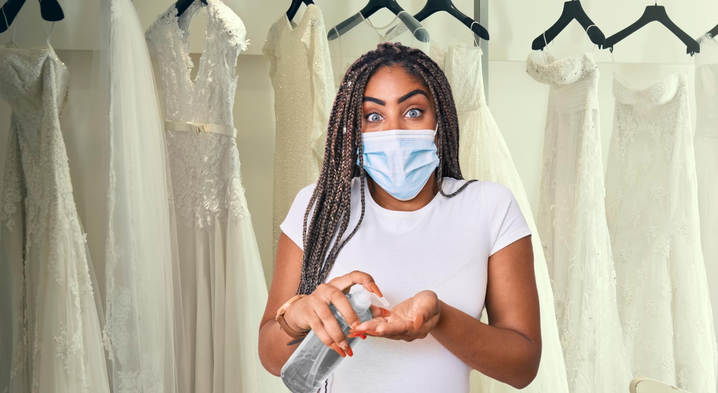 A Black woman wearing a medical mask and pumping sanitizer in her hand in front of several wedding dresses