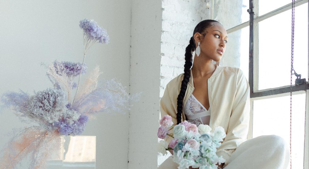 Black bride wearing a cream track suit looks outside of the window while holding a blue and purple bouquet.