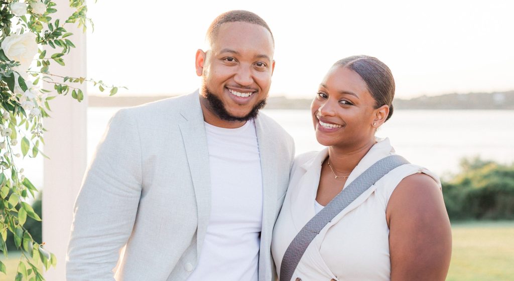 A smiling couple posing outdoors in soft natural light, with a scenic background of water and greenery. The man is wearing a light-colored blazer and white shirt, while the woman is dressed in an elegant sleeveless outfit, accented by floral greenery on the left side of the frame.