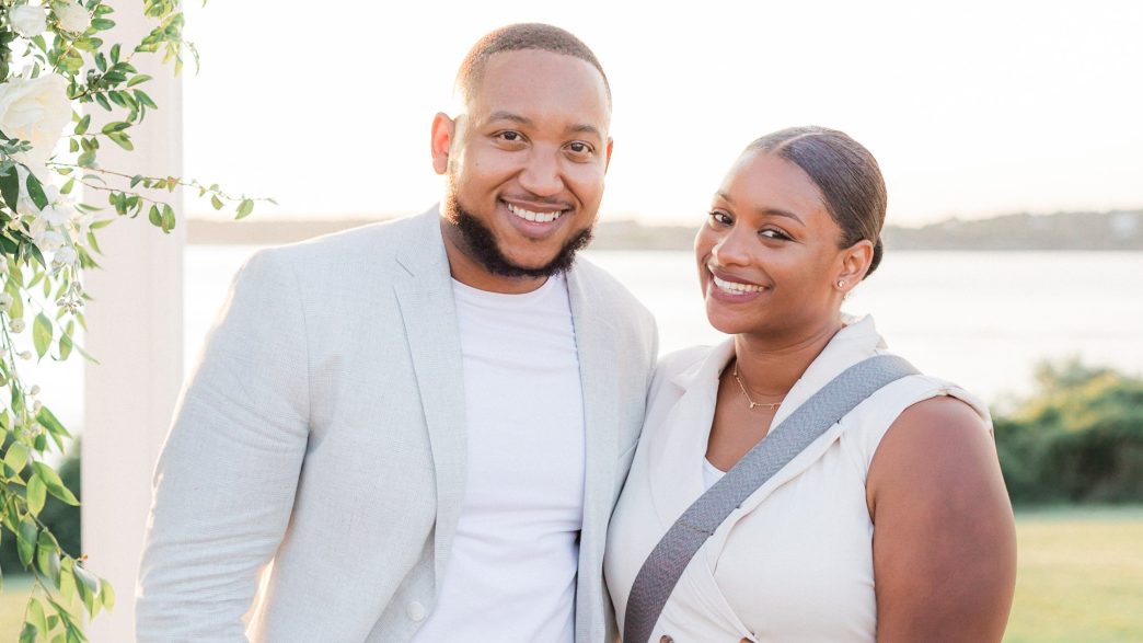 A smiling couple posing outdoors in soft natural light, with a scenic background of water and greenery. The man is wearing a light-colored blazer and white shirt, while the woman is dressed in an elegant sleeveless outfit, accented by floral greenery on the left side of the frame.