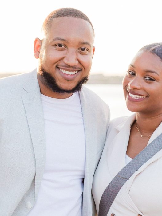 A smiling couple posing outdoors in soft natural light, with a scenic background of water and greenery. The man is wearing a light-colored blazer and white shirt, while the woman is dressed in an elegant sleeveless outfit, accented by floral greenery on the left side of the frame.