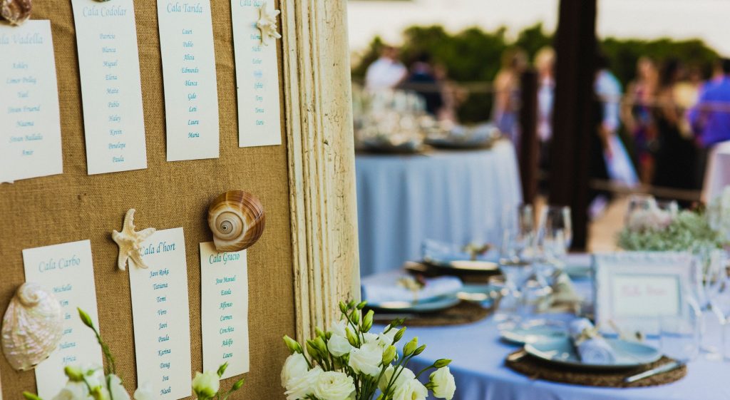 A close-up of a wedding seating chart displayed on a burlap-covered board, adorned with decorative seashells and starfish. Each section lists guest names under headings, with a blurred background showing an outdoor reception setup featuring round tables with blue tablecloths, elegant place settings, and glasses.