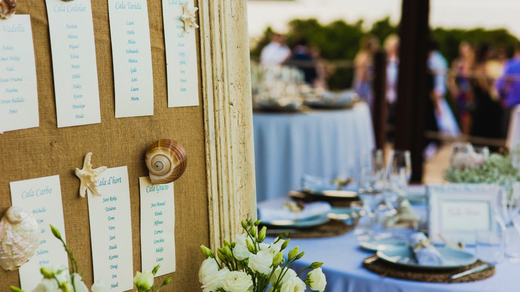 A close-up of a wedding seating chart displayed on a burlap-covered board, adorned with decorative seashells and starfish. Each section lists guest names under headings, with a blurred background showing an outdoor reception setup featuring round tables with blue tablecloths, elegant place settings, and glasses.