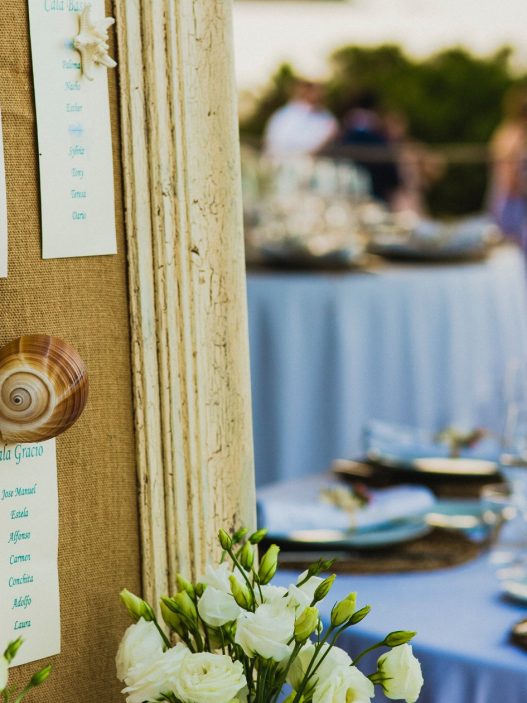 A close-up of a wedding seating chart displayed on a burlap-covered board, adorned with decorative seashells and starfish. Each section lists guest names under headings, with a blurred background showing an outdoor reception setup featuring round tables with blue tablecloths, elegant place settings, and glasses.