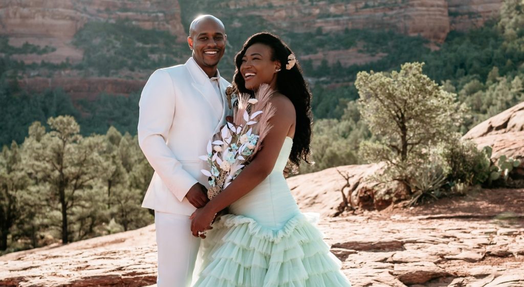 A person in a white tuxedo is next to a person in a wedding dress and they are both in front of a mountain