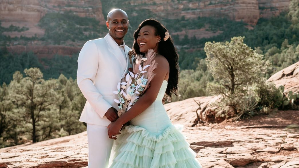 A person in a white tuxedo is next to a person in a wedding dress and they are both in front of a mountain