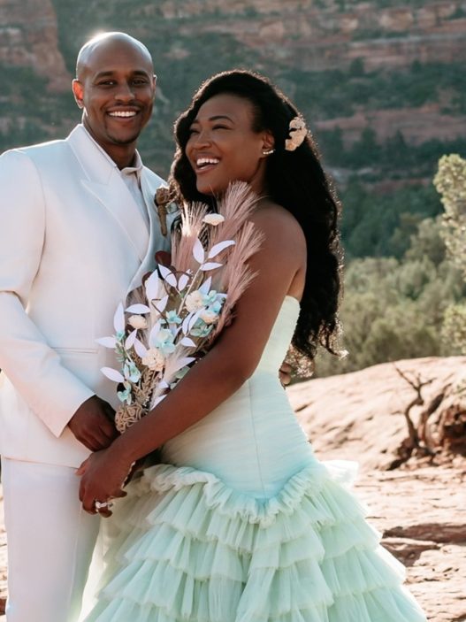 A person in a white tuxedo is next to a person in a wedding dress and they are both in front of a mountain