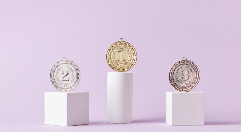 Three medals displayed on white podiums against a soft lavender background, with the gold medal labeled '1' on the tallest podium, the silver medal labeled '2' on a medium-height podium, and the bronze medal labeled '3' on the shortest podium.