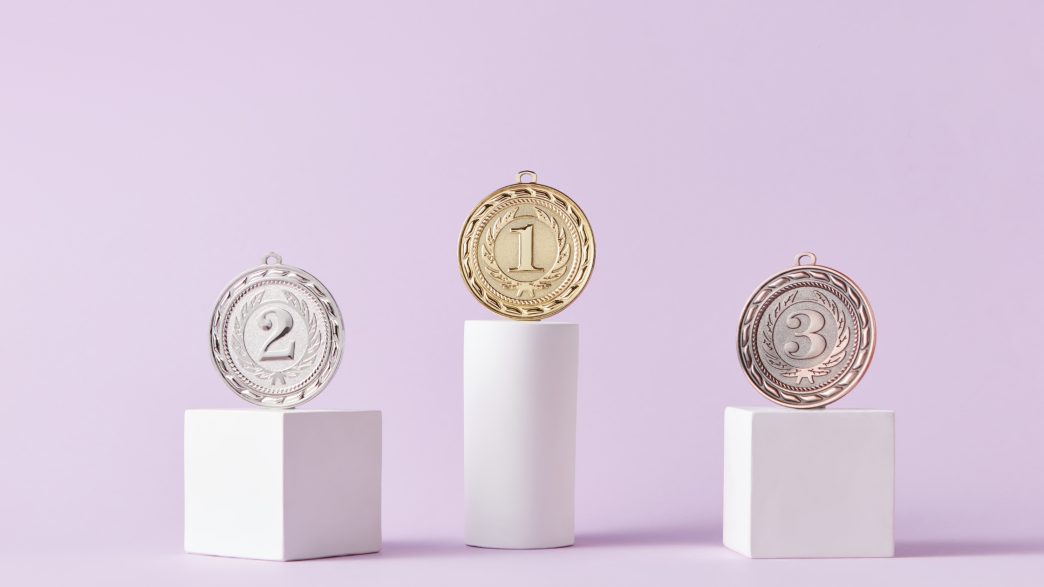 Three medals displayed on white podiums against a soft lavender background, with the gold medal labeled '1' on the tallest podium, the silver medal labeled '2' on a medium-height podium, and the bronze medal labeled '3' on the shortest podium.