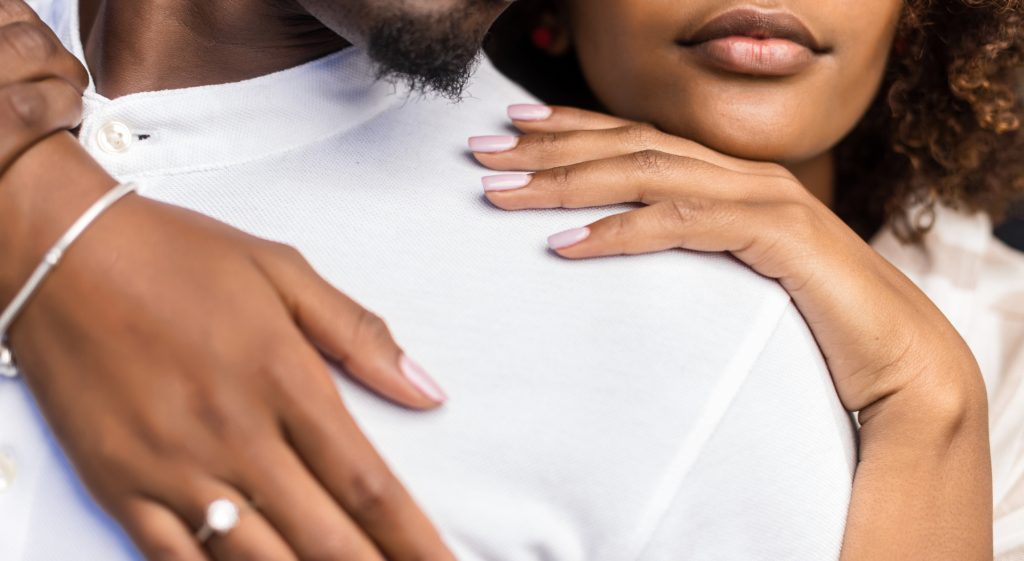 Close-up of a couple in an intimate embrace, with a focus on their hands resting gently on each other. The woman’s hand, adorned with a silver bracelet and an engagement ring, is tenderly placed on the man's shoulder, both exuding warmth and connection.