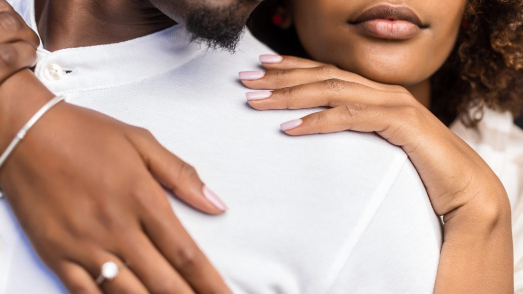 Close-up of a couple in an intimate embrace, with a focus on their hands resting gently on each other. The woman’s hand, adorned with a silver bracelet and an engagement ring, is tenderly placed on the man's shoulder, both exuding warmth and connection.