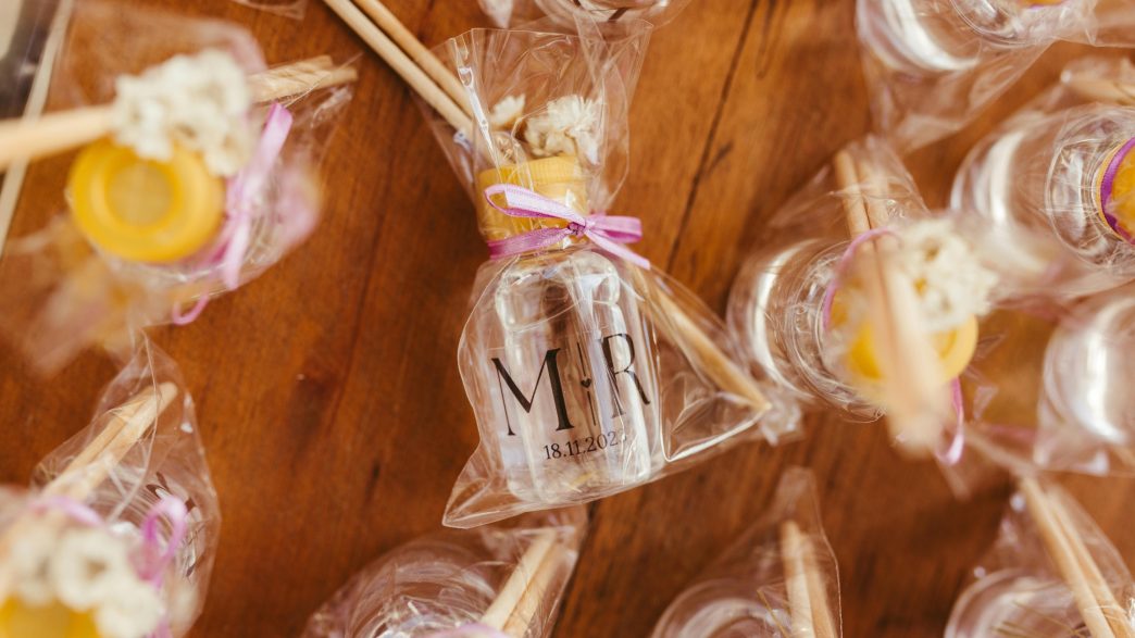 Close-up of wedding favors wrapped in clear cellophane, featuring small bottles labeled with 'M ♥ R' and a date, tied with pink ribbons. The packages include honey sticks and dried flowers, displayed on a wooden table.