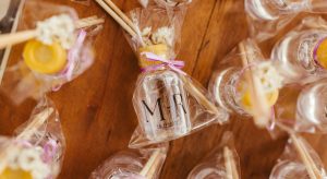 Close-up of wedding favors wrapped in clear cellophane, featuring small bottles labeled with 'M ♥ R' and a date, tied with pink ribbons. The packages include honey sticks and dried flowers, displayed on a wooden table.