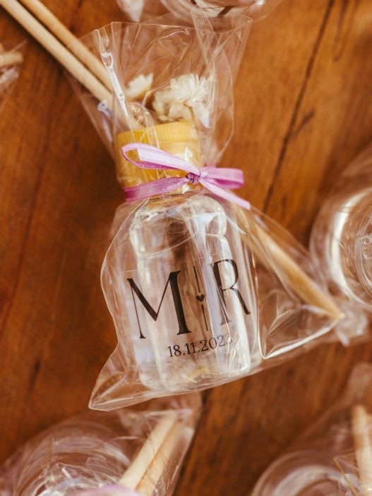 Close-up of wedding favors wrapped in clear cellophane, featuring small bottles labeled with 'M ♥ R' and a date, tied with pink ribbons. The packages include honey sticks and dried flowers, displayed on a wooden table.