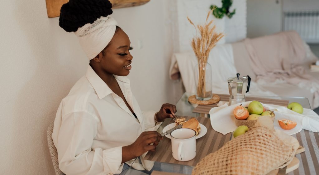 A serene scene of a woman sitting at a table enjoying a meal, wearing a white headwrap and shirt. The table is set with a plate of food, a mug, fresh fruit, and a moka pot, while the background features cozy home decor, including a vase with dried wheat and a soft beige couch.