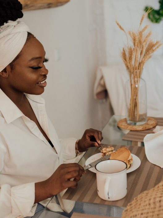 A serene scene of a woman sitting at a table enjoying a meal, wearing a white headwrap and shirt. The table is set with a plate of food, a mug, fresh fruit, and a moka pot, while the background features cozy home decor, including a vase with dried wheat and a soft beige couch.