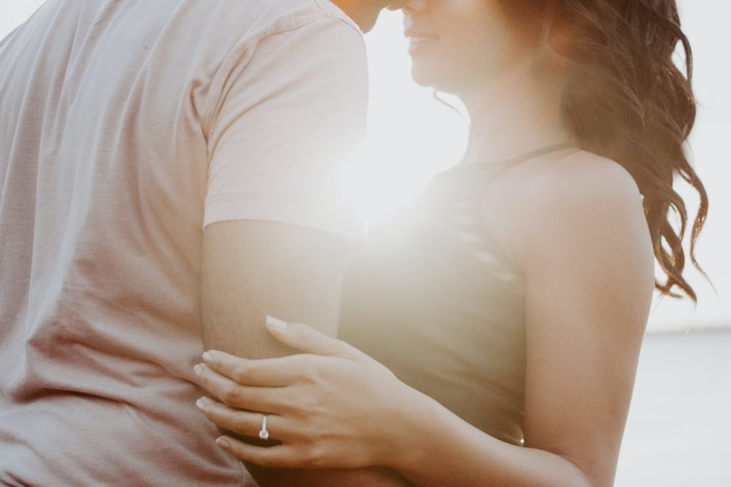 A close-up of a couple embracing at sunset, with the focus on the woman's hand resting on the man's arm, showcasing her engagement ring.