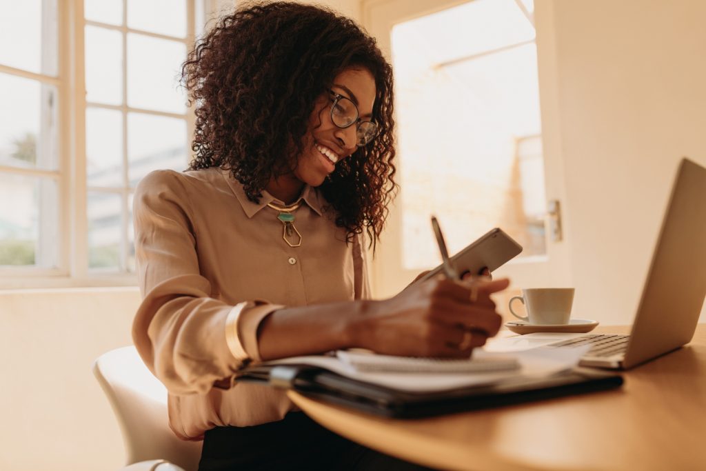 A Black woman is smiling as she sits at a table with a laptop