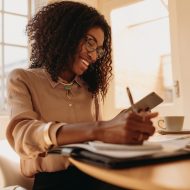 A Black woman is smiling as she sits at a table with a laptop