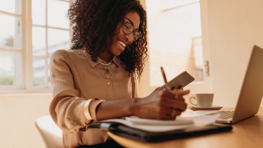 A Black woman is smiling as she sits at a table with a laptop