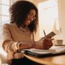 A Black woman is smiling as she sits at a table with a laptop