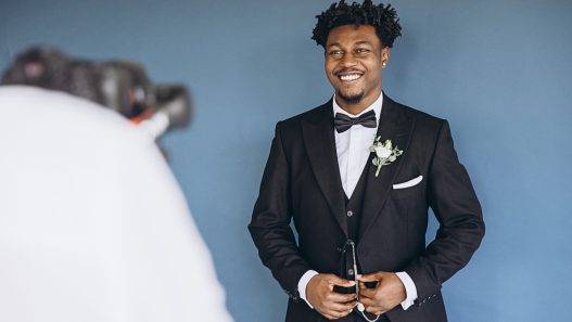 A Black groom in a tuxedo and bow tie smiles at the camera.