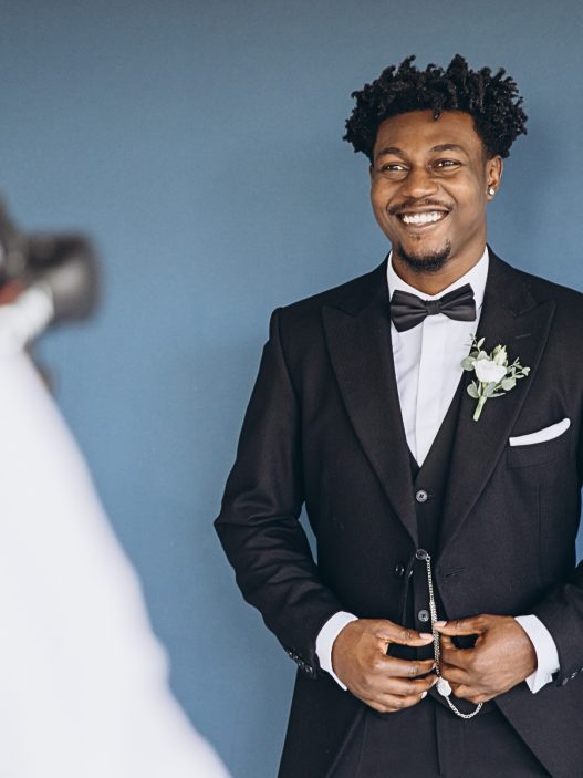 A Black groom in a tuxedo and bow tie smiles at the camera.