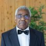 An older Black man who is the father of the bride wearing a tuxedo stares emotionless at the camera