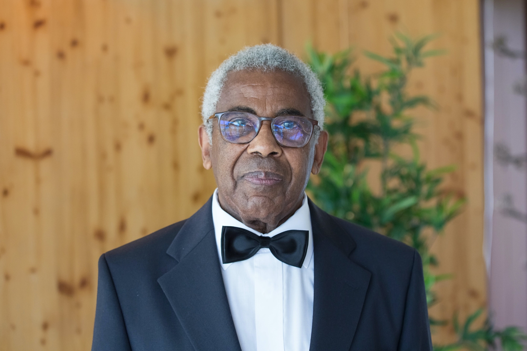 An older Black man who is the father of the bride wearing a tuxedo stares emotionless at the camera