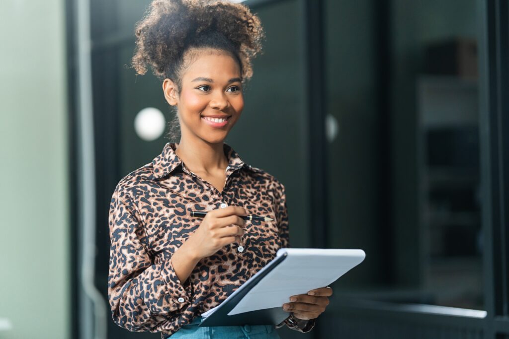 A smiling person in a leopard print shirt holds a pen and a notepad, looking to the side.