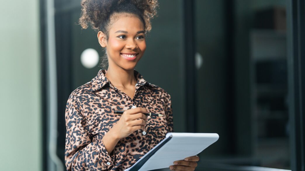 A smiling person in a leopard print shirt holds a pen and a notepad, looking to the side.