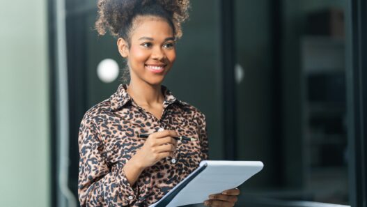 A smiling person in a leopard print shirt holds a pen and a notepad, looking to the side.