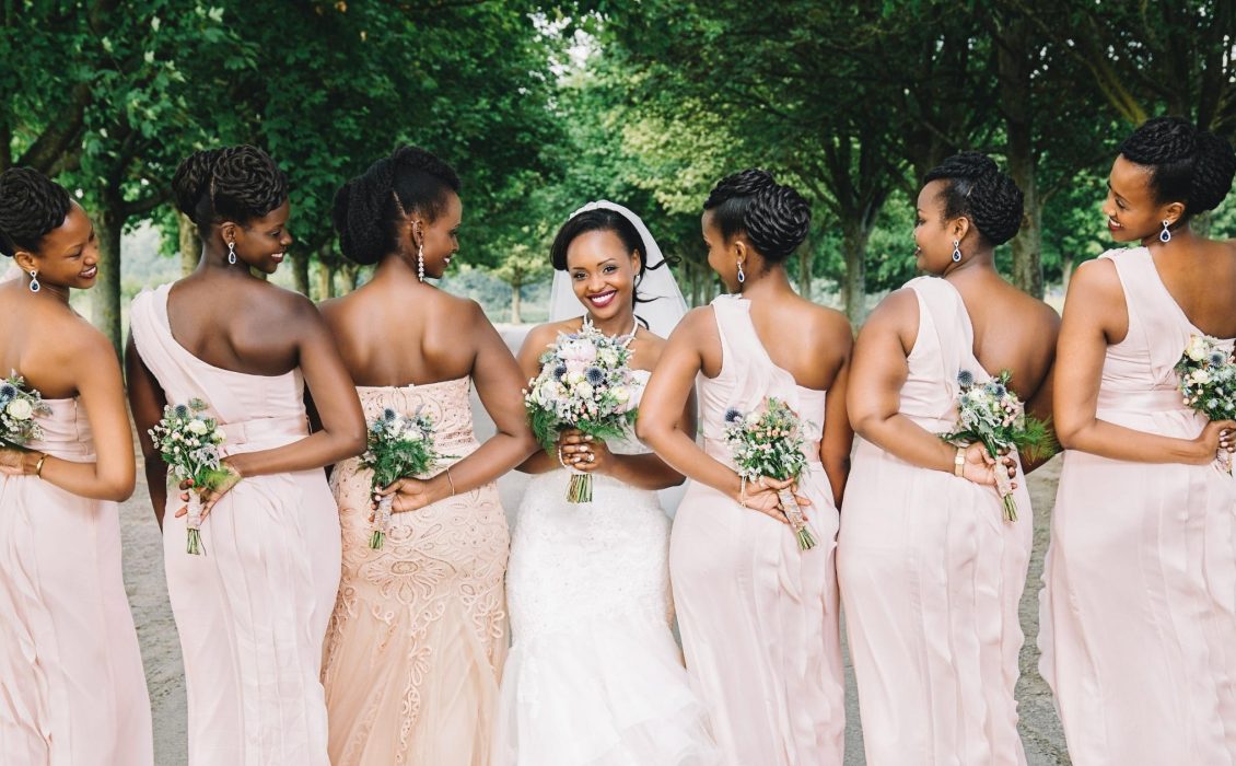 Six Black women in pink dresses stand next to a Black woman in a white dress
