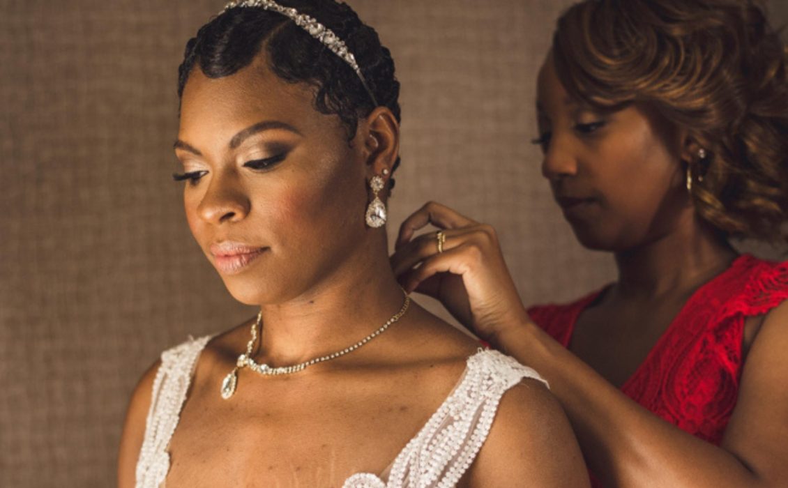 A Black bride is standing as someone helps her with a necklace