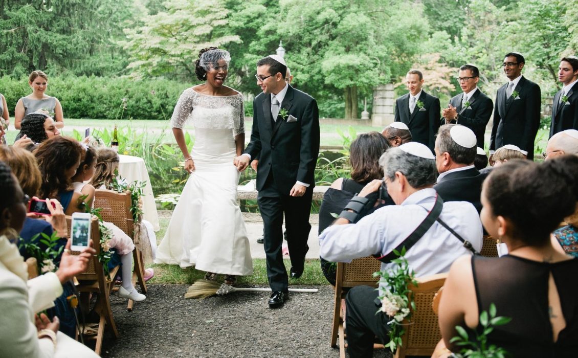 A Black woman and a Jewish man jump over a broom in front of people sitting down