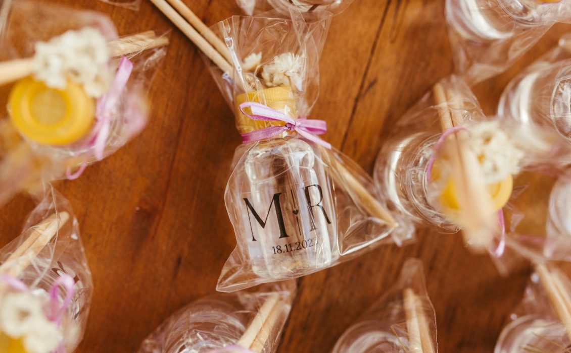 Close-up of wedding favors wrapped in clear cellophane, featuring small bottles labeled with 'M ♥ R' and a date, tied with pink ribbons. The packages include honey sticks and dried flowers, displayed on a wooden table.