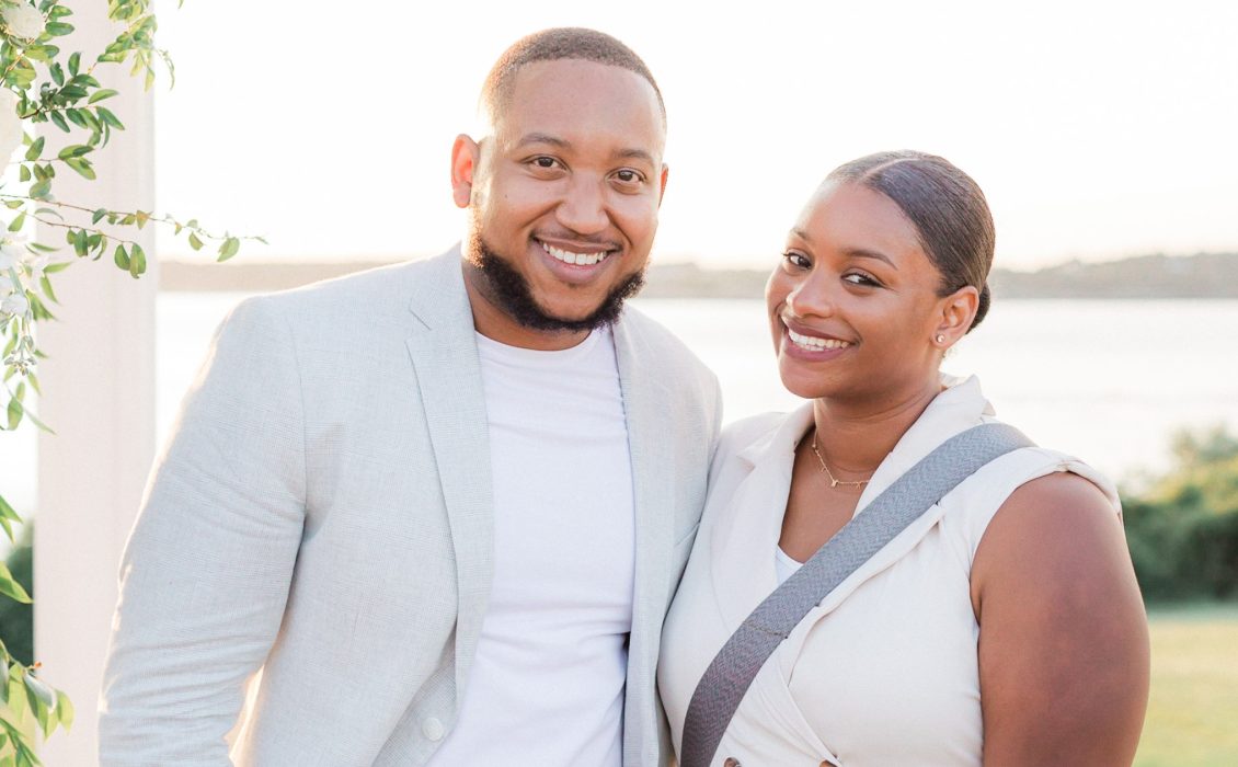 A smiling couple posing outdoors in soft natural light, with a scenic background of water and greenery. The man is wearing a light-colored blazer and white shirt, while the woman is dressed in an elegant sleeveless outfit, accented by floral greenery on the left side of the frame.